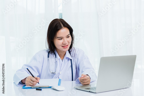 Asian doctor young beautiful woman smiling using working with a laptop computer and her writing something on paperwork or clipboard white paper at hospital desk office, Healthcare medical concept © sorapop