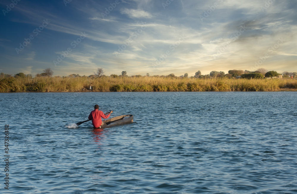 One young African adult wearing a bright red shirt in a handmade wooden boat on the Kavango River in Namibia, Africa One young African adult wearing a bright red shirt in a handmade wooden boat on the Kavango River in Namibia, Africa
