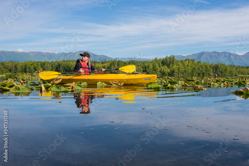 Young teen boy paddles canoe on lily pad-covered lake in Alaska with blue sky and mountains in the background. 