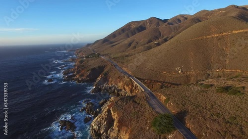 Aerial: Vehicles moving over coastal road on mountains by rocky coastline against sky, waves splashing in sea during sunset - Big Sur, California photo