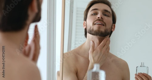Smiling confident handsome young guy applying aftershave perfume lotion on face. Happy sexy shirtless bearded man putting fragrance on neck skin after shaving looking in mirror. Men grooming concept. photo