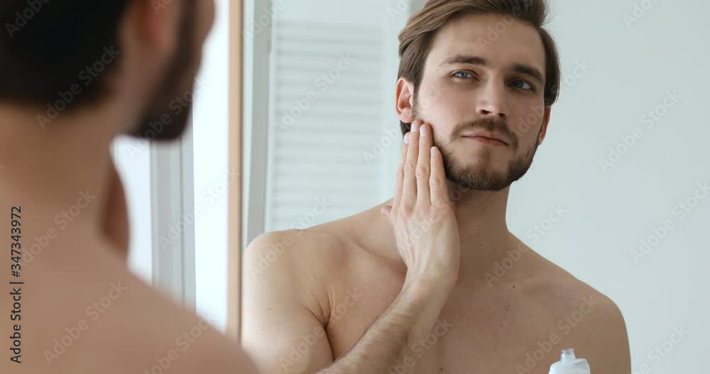 Confident bearded sexy shirtless man holding lotion bottle applying moisturizing balm on face and neck after shaving looking in bathroom mirror. Aftershave skin care treatment concept. Close up view Confident bearded sexy shirtless man holding lotion bottle applying moisturizing balm on face and neck after shaving looking in bathroom mirror. Aftershave skin care treatment concept. Close up view