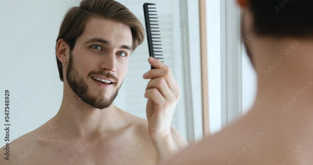 Confident attractive young adult shirtless man holding comb brushing hair looking in bathroom mirror. Smiling handsome bearded guy doing hairstyle in the morning. Men beauty hair care concept.