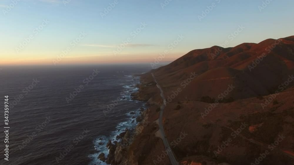 Aerial: Drone moving over waves splashing on rocky coastline in sea by coastal road against sky during sunset - Big Sur, California Aerial: Drone moving over waves splashing on rocky coastline in sea by coastal road against sky during sunset - Big Sur, California