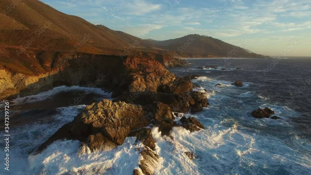 Aerial: Drone approaching rocky coastline while waves splashing in sea at beach during sunset, scenic view of mountains against sky - Big Sur, California