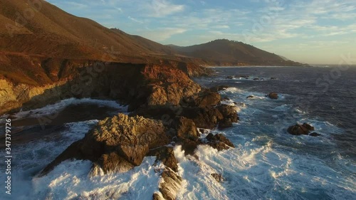 Aerial: Drone approaching rocky coastline while waves splashing in sea at beach during sunset, scenic view of mountains against sky - Big Sur, California