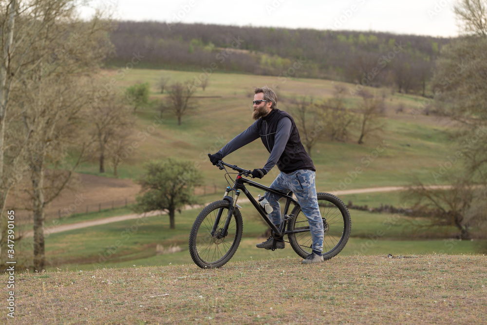 Sports brutal bearded guy on a modern mountain bike. Cyclist on the green hills in the spring.