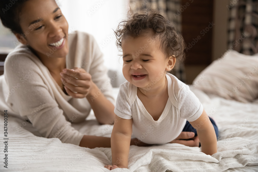 Happy young african American mother relax play on bed at home with little baby infant, cute small toddler child go on hands and knees, have fun with biracial mom in bedroom, childcare concept Happy young african American mother relax play on bed at home with little baby infant, cute small toddler child go on hands and knees, have fun with biracial mom in bedroom, childcare concept