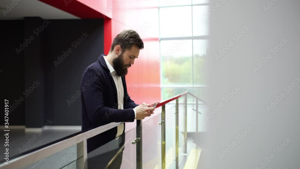 Close-up side view of calm bearded man typing sms on the phone leaning on glass fence inside modern office building with red walls and panoramic windows. Brunette guy is texting message while standing Close-up side view of calm bearded man typing sms on the phone leaning on glass fence inside modern office building with red walls and panoramic windows. Brunette guy is texting message while standing