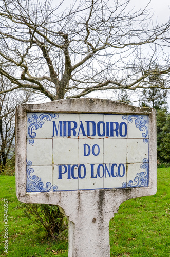 Blue sign Miradoiro do Pico Longo, Viewpoint Pico Longo in English, on typical Portuguese tiles. Grass and trees without leaves in the background. Vertical photo.