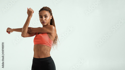 Fototapeta Naklejka Na Ścianę i Meble -  Sporty girl. Beautiful young african woman in sportswear stretching her arms and looking at camera while standing against grey background