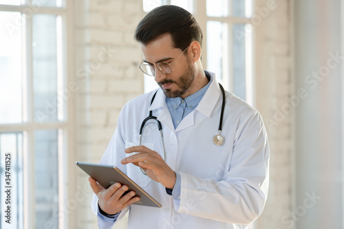 Serious young Caucasian male doctor in medical uniform and stethoscope fill patient anamnesis or history on modern tablet, man GP or physician consult client online using pad, healthcare concept