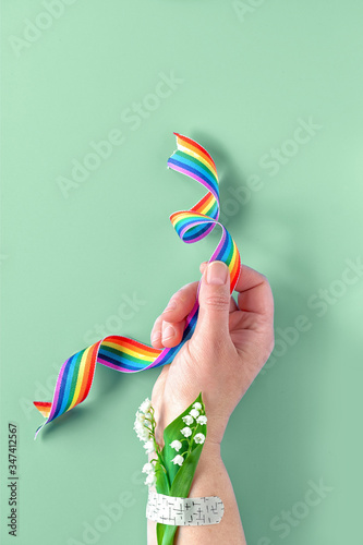 Rainbow ribbon in hand. Lily of the valley flowers, small bouquet attached to wrist with medical patch, aid band. Thank you doctors, nurses and key workers! Creative top view on green background.