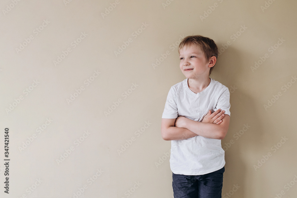 A happy boy of European appearance in a white T-shirt looks into the distance. Photo of a 7 years old baby on a beige background. A happy boy of European appearance in a white T-shirt looks into the distance. Photo of a 7 years old baby on a beige background.