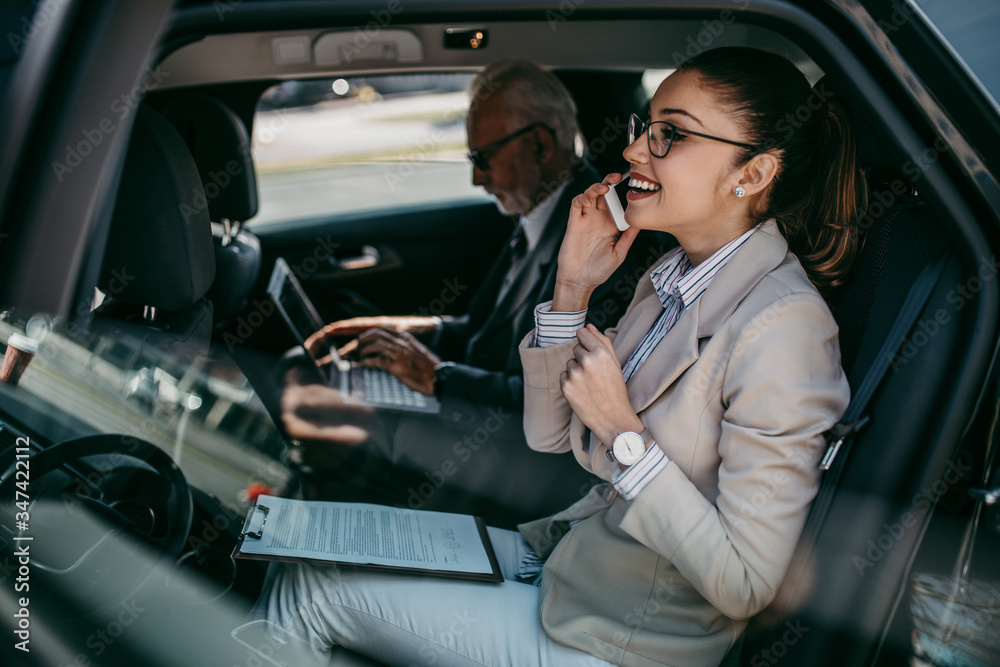 Good looking senior business man and his young woman colleague or coworker sitting on backseat in luxury car. They talking, smiling and using laptop and smart phones. Good looking senior business man and his young woman colleague or coworker sitting on backseat in luxury car. They talking, smiling and using laptop and smart phones.