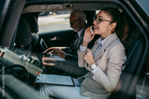 Good looking senior business man and his young woman colleague or coworker sitting on backseat in luxury car. They talking, smiling and using laptop and smart phones. 