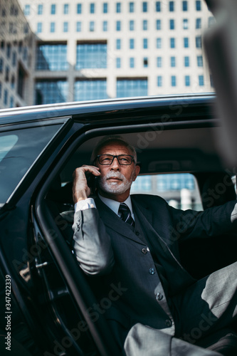 Good looking senior business man sitting on backseat in luxury car. He opens car doors and going or stepping out. Big business building in background. Transportation in corporate business concept. © Dusko