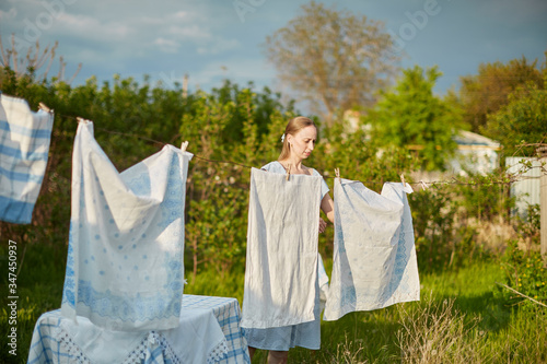 Blonde woman villager hanging wet white-blue laundry on clothesline to dry in the backyard. A green tree in the background.