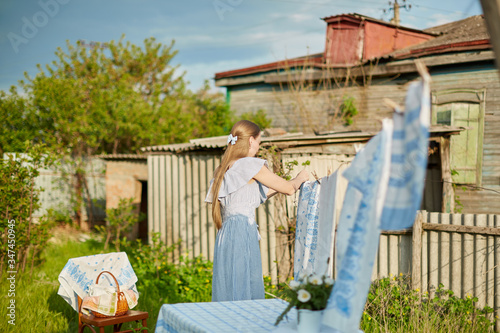 Blonde woman villager hanging wet white-blue laundry on clothesline to dry in the backyard. A wooden old house in the background.