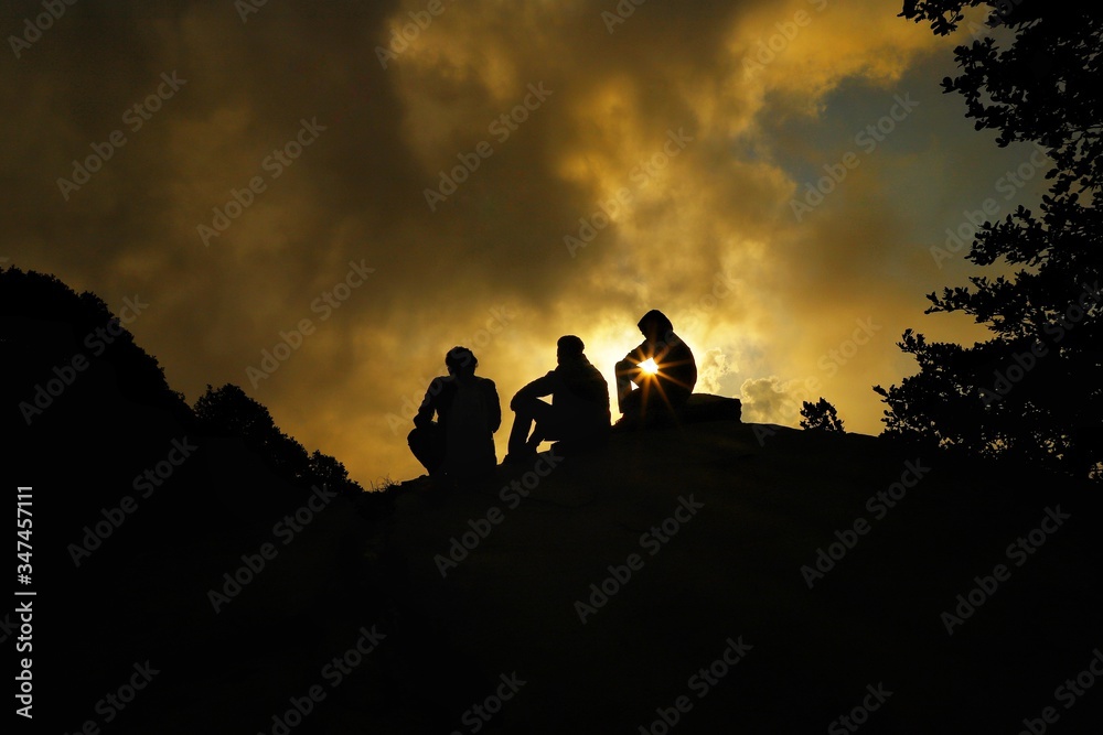 the thre man is sitting  on top of a mountain as the sun sets at hatu peak,himachal  pradesh,india.