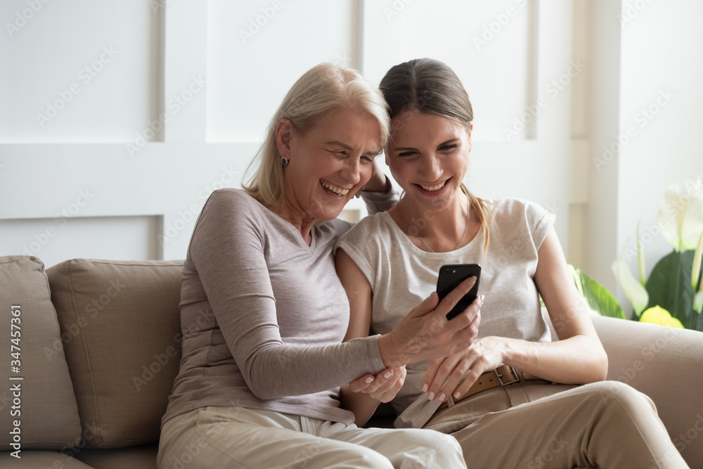 Excited Middle Aged Mom And Grownup Daughter Sit On Couch At Home Laugh Watching Funny Video On
