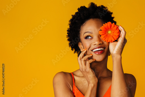 Portrait of a happy excited young woman holding orange gerbera daisy covering her eye and looking up at copy space