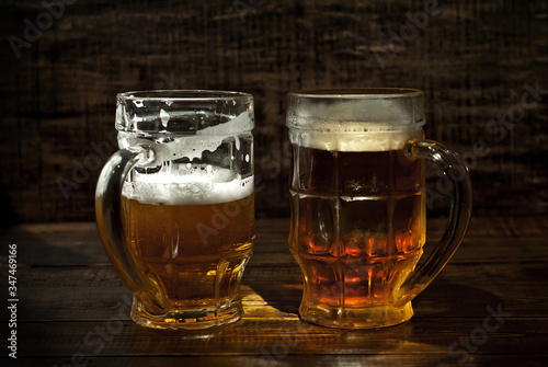 A glass of amber beer. Glasses on a wooden table. Black shabby boards on the background. Bright contrasting light and glare on the glass.