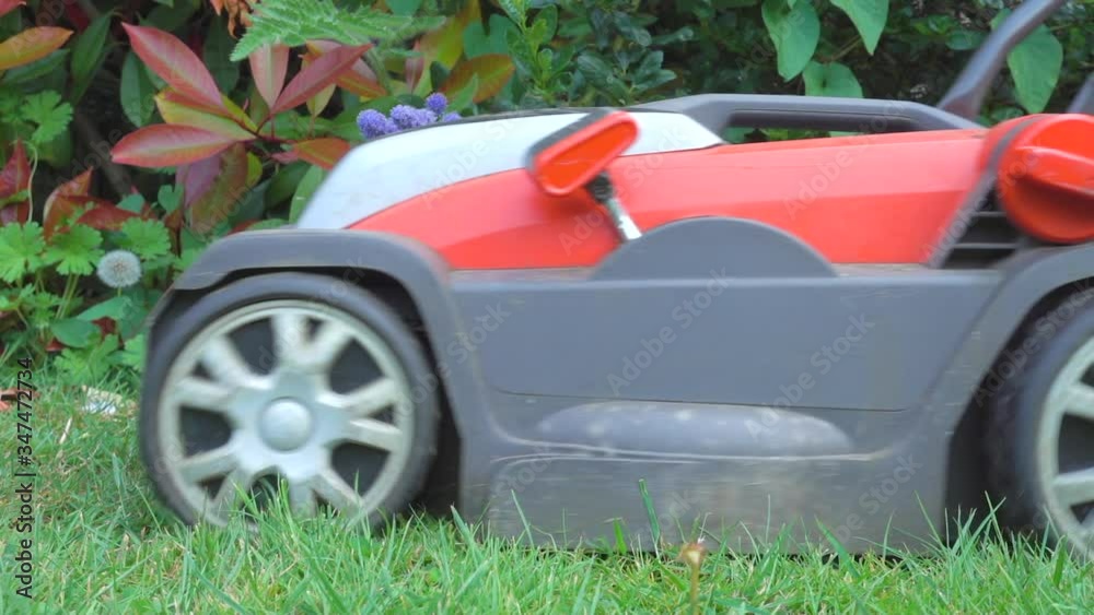 Close POV slow motion shot of a modern, lightweight, cordless (battery operated) lawn mower, being pushed over grass past garden / yard bushes. Close POV slow motion shot of a modern, lightweight, cordless (battery operated) lawn mower, being pushed over grass past garden / yard bushes.