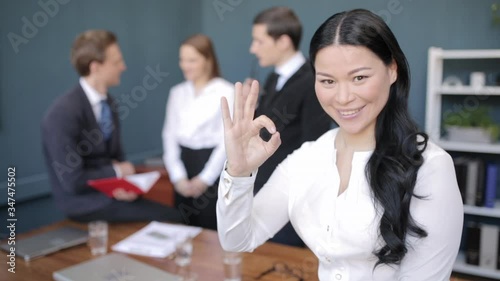 Asian business lady smiles at the camera and shows OK symbol, everything is fine on the background of male businessmen in business clothes standing in the board or conferece room. Prores 422 photo