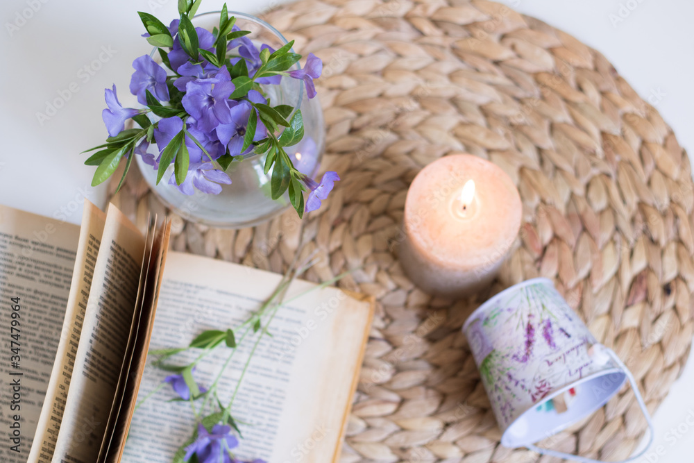 An open book in the foreground with a periwinkle flower between pages. Vase with flowers. Burning candle. An open book in the foreground with a periwinkle flower between pages. Vase with flowers. Burning candle.