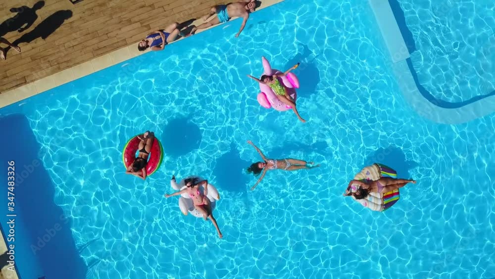 Aerial. Friends chilling in swimming pool with inflatable flamingo, swan, mattress. Happy young people bathe on floating mattresses in luxury resort. View from above. Girls in bikini sunbathing in sun
