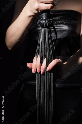 BDSM concept. A woman in a leather dress holds a black whip with a long fringe. Role-playing games for adults. Close-up of sex toys for domination. © Михаил Решетников