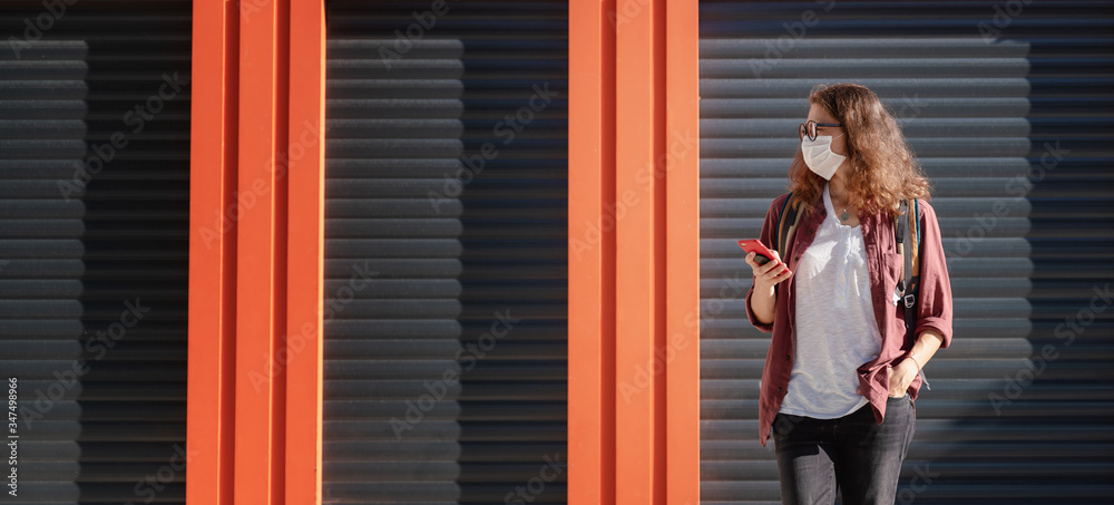 Fototapeta premium Attractive serious young woman in a mask and eyeglasses looks at the screen on a smartphone, looking for information. Against the backdrop of a closed cafe and shop during the coronavirus pandemic.