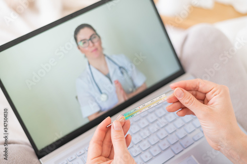 A woman is lying in bed with heat and is holding a thermometer. A friendly doctor talks to a sick patient by video link. Therapist on a laptop screen. Online medical consultation during the quarantine