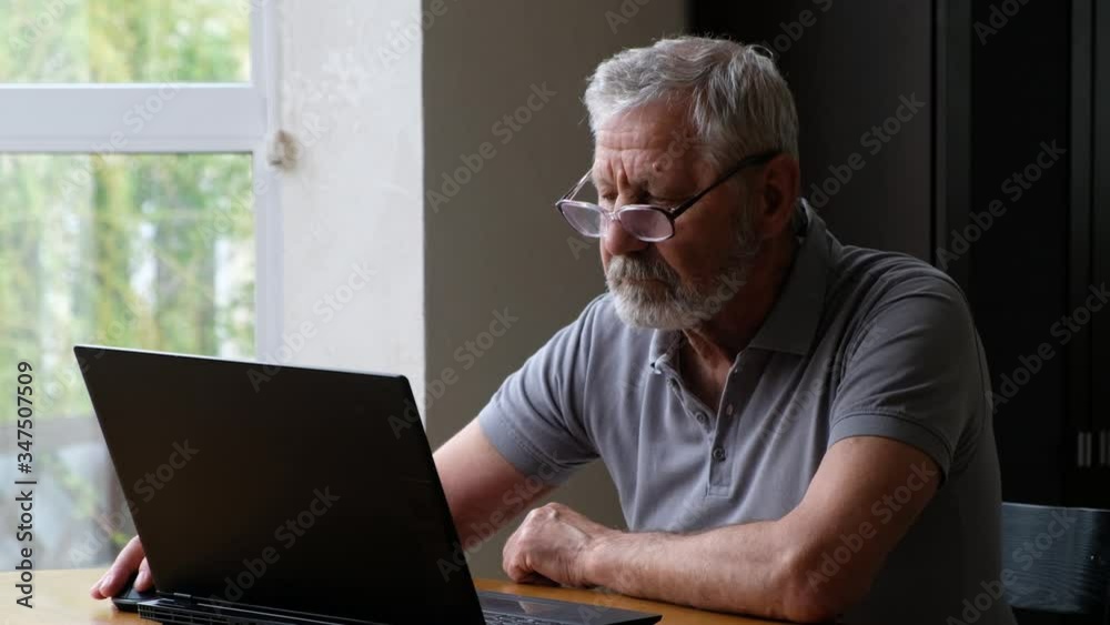Senoir elderly man in glasses sitting at the table and looking at laptop Senoir elderly man in glasses sitting at the table and looking at laptop