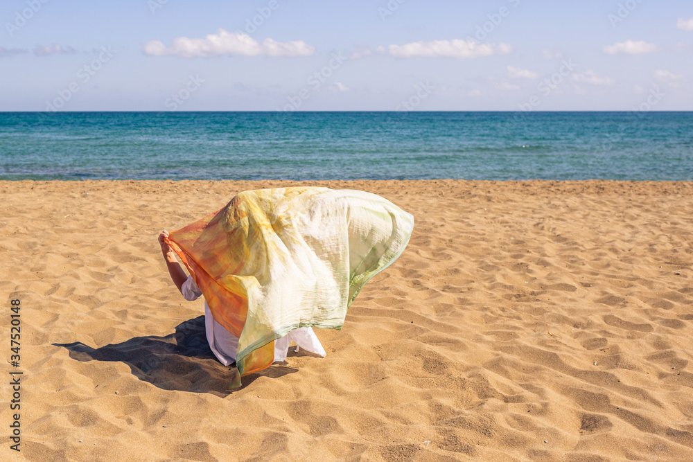 Back view of lonely Muslim woman traveler in a colorful headscarf, scarf sitting on the sand on the beach and looking on the sea. Back view of lonely Muslim woman traveler in a colorful headscarf, scarf sitting on the sand on the beach and looking on the sea.