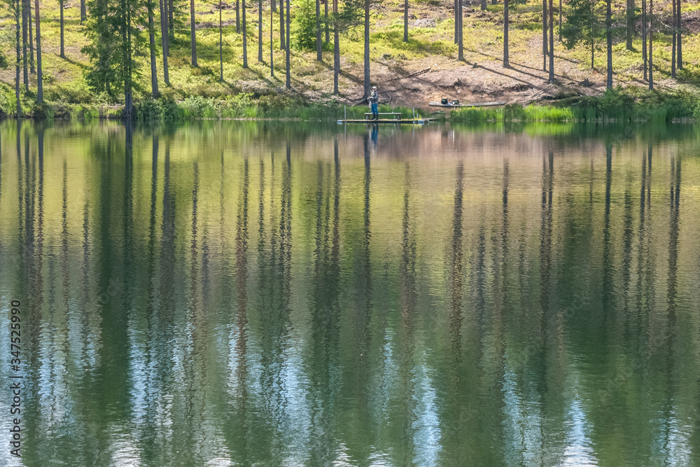 sky and tree reflection in secluded area by the lake. Calm waters and cloudy sky. fisherman in the background Sweden. selective focus. long exposure sky and tree reflection in secluded area by the lake. Calm waters and cloudy sky. fisherman in the background Sweden. selective focus. long exposure