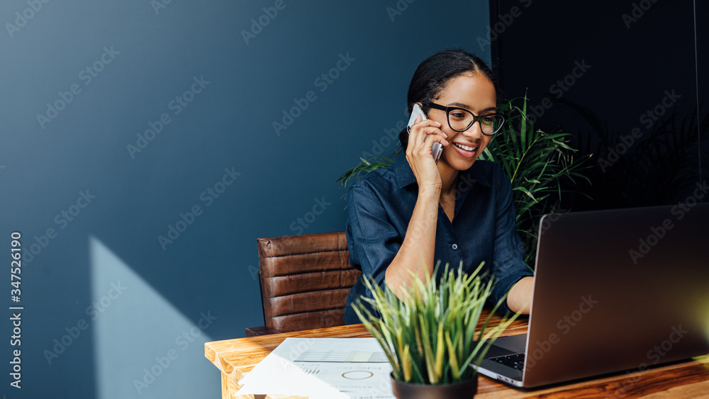 Entrepreneur sitting at table typing on a laptop while talking on cell phone  - obrazy, fototapety, plakaty 