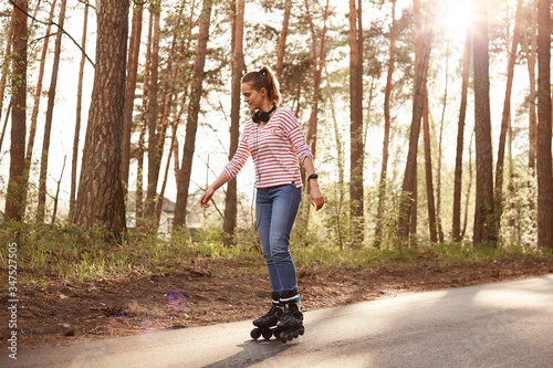 Full length photo of adorable female with ponytail and headphones around beck, woman dresses striped shirt and jeans, listening to music, enjoying songs and beautiful nature. Lifestyle concept. photo