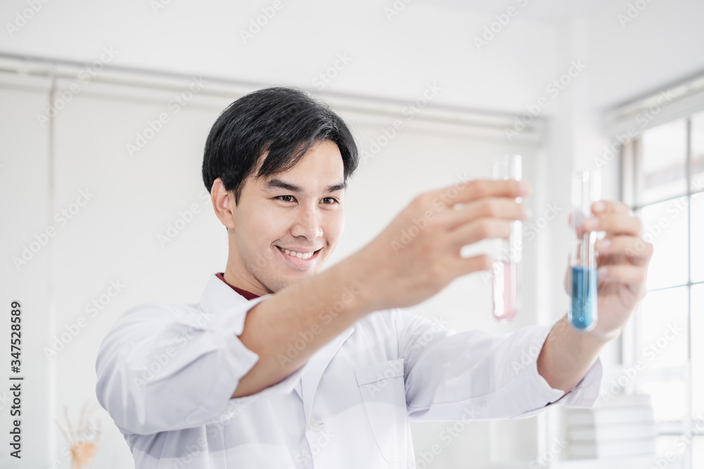 A young male scientist with black hair wearing white lab coat smiling and holding up test tubes with blue and pink solution in a laboratory setting. A young male scientist with black hair wearing white lab coat smiling and holding up test tubes with blue and pink solution in a laboratory setting.