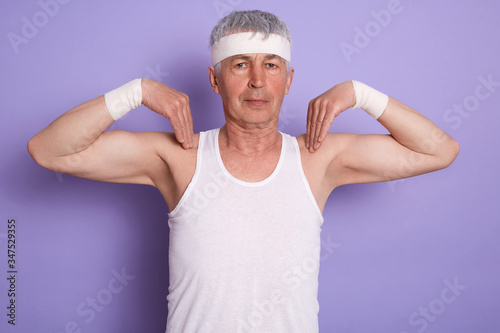 Image of concentrated energetic man standing isolated over lilac background in studio, raising arms, doing physical exercises, being fond of sports, having training, getting results. Sport concept.