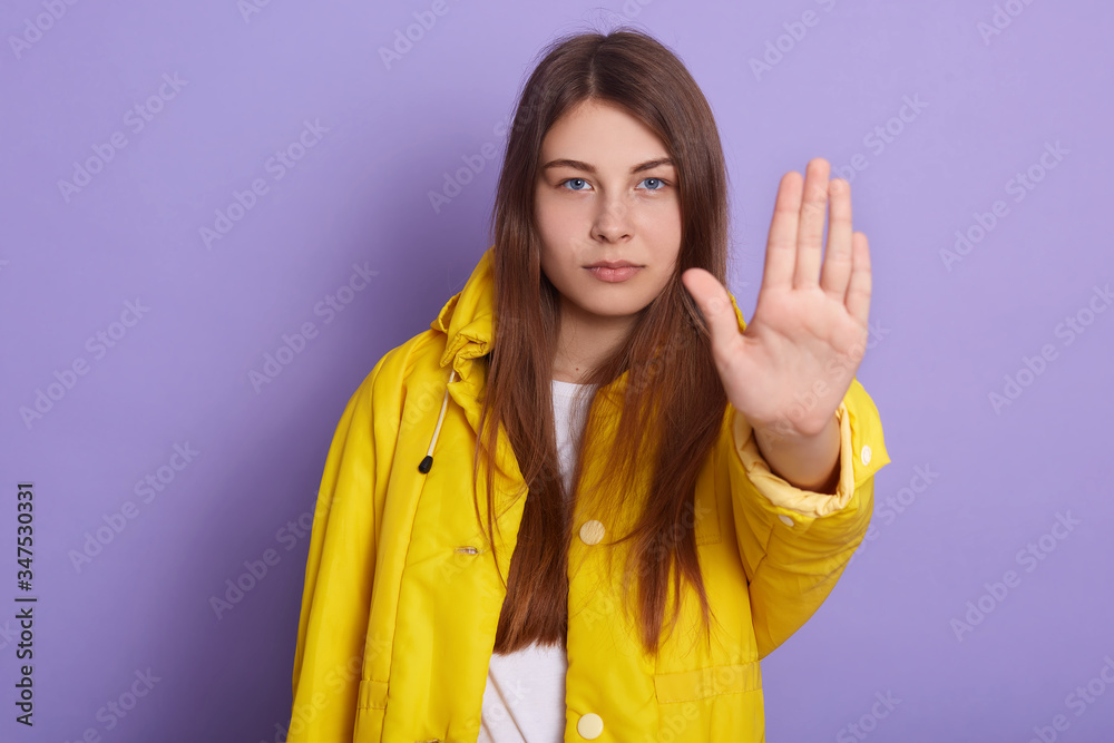 Image of serious woman showing stop gesture with her palm, wearing yellow jacket and white shirt, posing isolated over lilac studio background, winsome female with long hair. People concept. Image of serious woman showing stop gesture with her palm, wearing yellow jacket and white shirt, posing isolated over lilac studio background, winsome female with long hair. People concept.