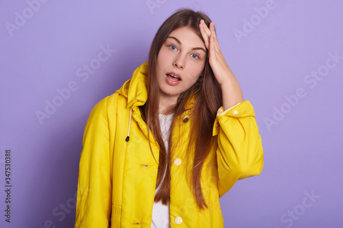 Studio shot of teenage girl with opened mouth and hands on head, forgot something important, remembering, wearing yellow jacket, posing isolated over lilac studio background.People emotions concept. © sementsova321