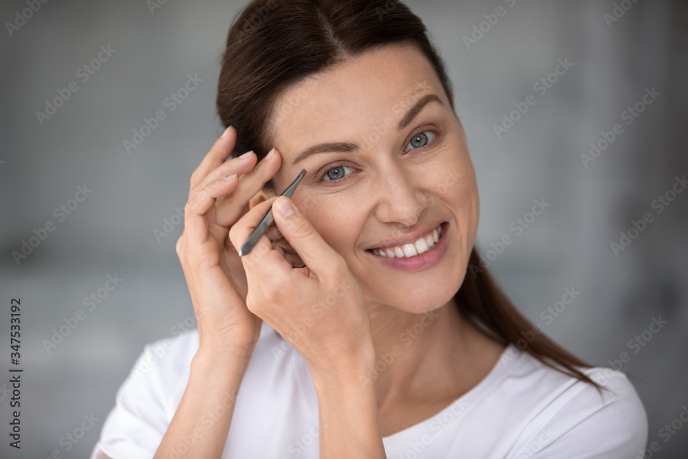 Close up portrait of attractive 30s caucasian woman looking at camera holding tweezers pluck eyebrows, makes perfect arch takes care of herself, beauty salon at home, routine, easy to use tool concept Close up portrait of attractive 30s caucasian woman looking at camera holding tweezers pluck eyebrows, makes perfect arch takes care of herself, beauty salon at home, routine, easy to use tool concept