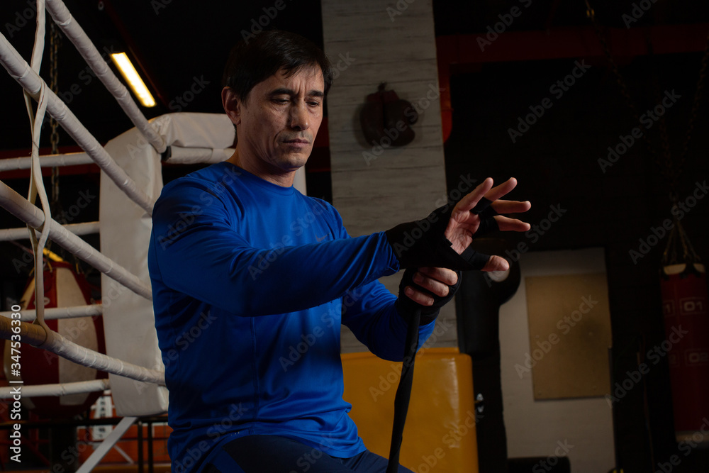  An experienced coach prepares for training in the boxing gym. Winds his hands with black bandages. 