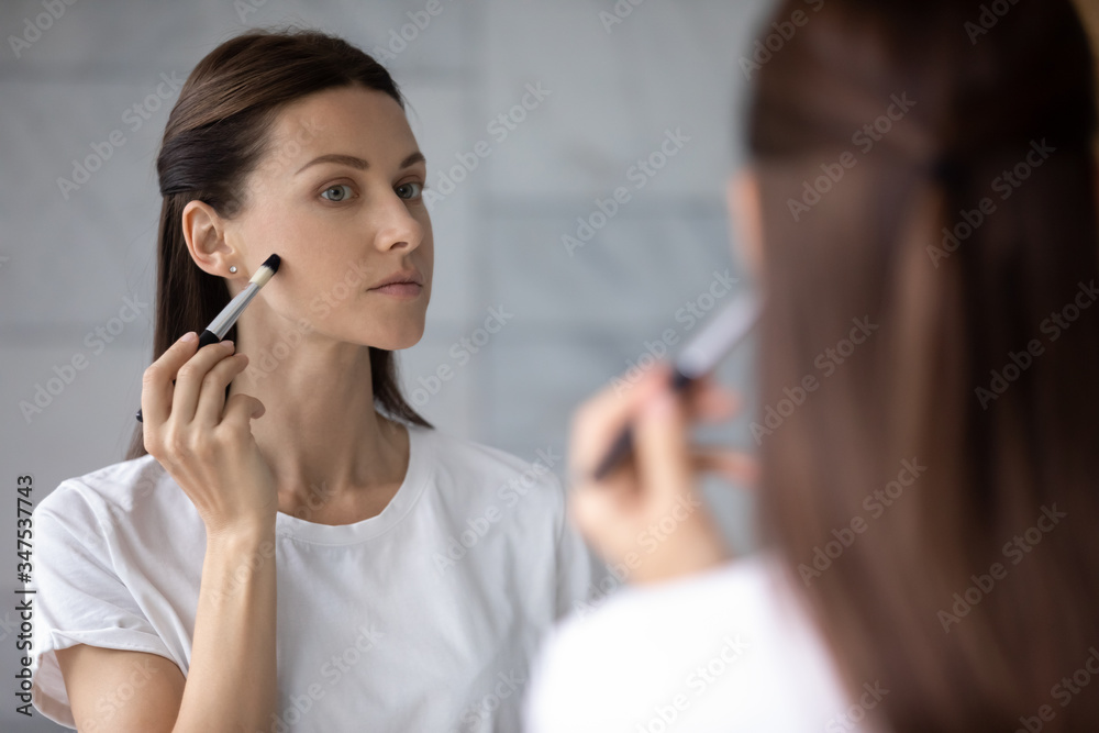 Over shoulder view serious pretty 35s woman reflected in mirror hold brush applying make up foundation concealer cream on perfect fresh skin. Grooming, morning routine, makeup, skincare beauty concept Over shoulder view serious pretty 35s woman reflected in mirror hold brush applying make up foundation concealer cream on perfect fresh skin. Grooming, morning routine, makeup, skincare beauty concept