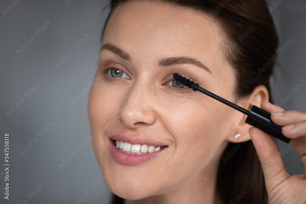 Close up face beautiful female gray-eyed 30s woman holding tube applying black mascara makes eyelashes more lengthening volumizing or thickening, look at mirror grooming herself before dating concept