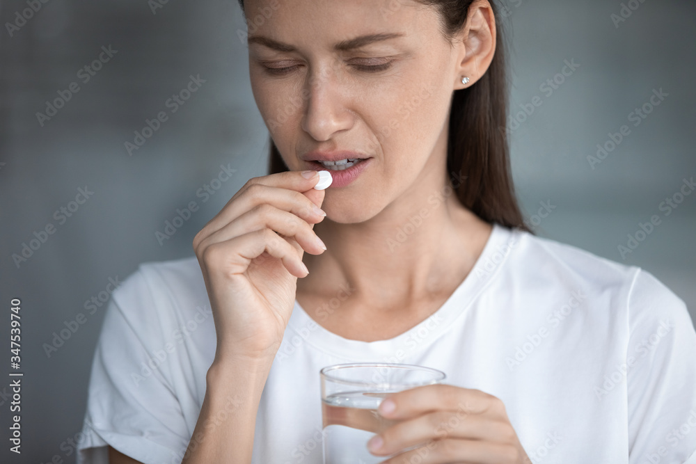 Close up image of unhealthy 35s female closed eyes suffers from migraine or hangover holds glass of water takes painkiller pill for throbbing headache reducing, medical abortion hard decision concept Close up image of unhealthy 35s female closed eyes suffers from migraine or hangover holds glass of water takes painkiller pill for throbbing headache reducing, medical abortion hard decision concept