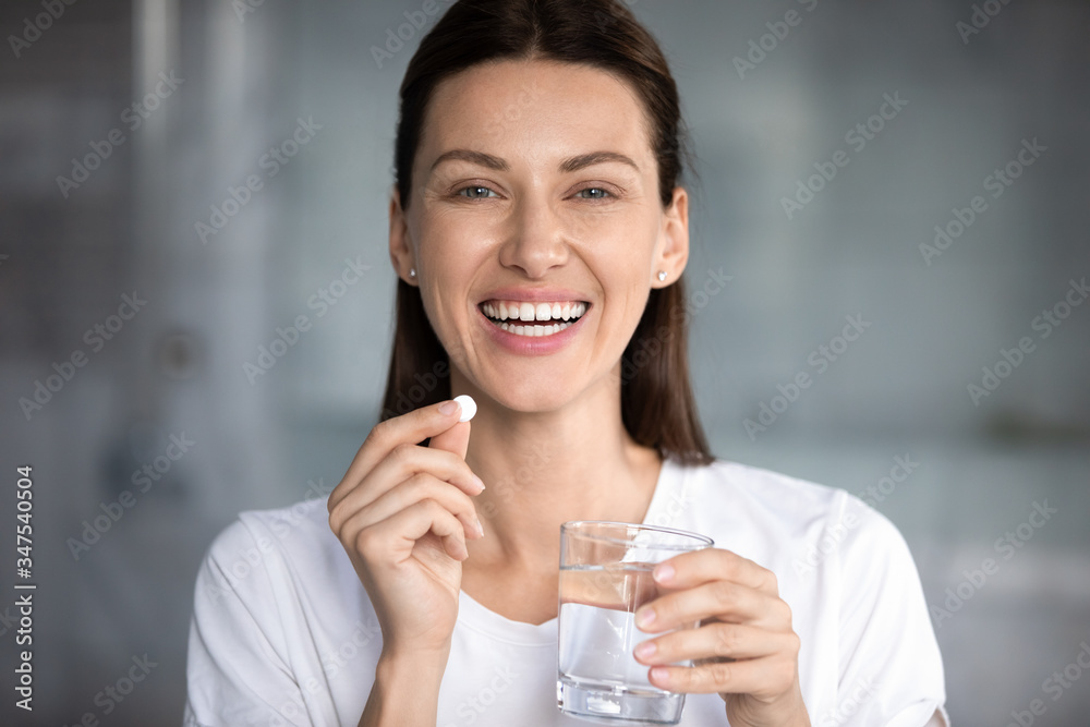 Head shot portrait happy woman holds pill glass of water looks at camera, takes daily medicine vitamin D, omega 3 supplements, skin hair nail strengthen and beauty, medication for health care concept