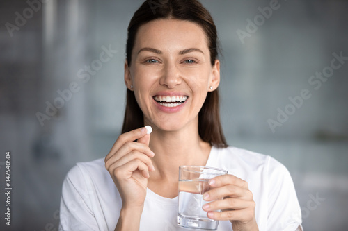 Head shot portrait happy woman holds pill glass of water looks at camera  takes daily medicine vitamin D  omega 3 supplements  skin hair nail strengthen and beauty  medication for health care concept